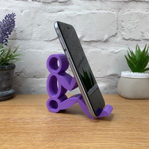 May include: Purple 3D printed phone stand shaped like a person holding a phone. The stand is on a wooden surface.