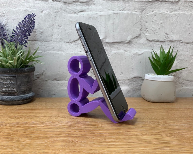 May include: Purple 3D printed phone stand shaped like a person holding a phone. The stand is on a wooden surface.