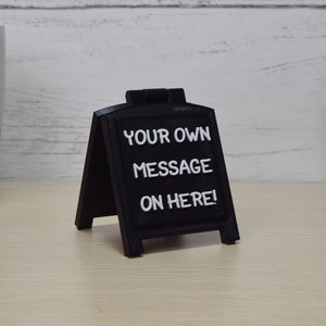 May include: A small, black chalkboard-style sign with white lettering that reads "YOUR OWN MESSAGE ON HERE!". The sign is in an A-frame style, suitable for tabletop display. The background includes a white and gold mug and a small potted plant.