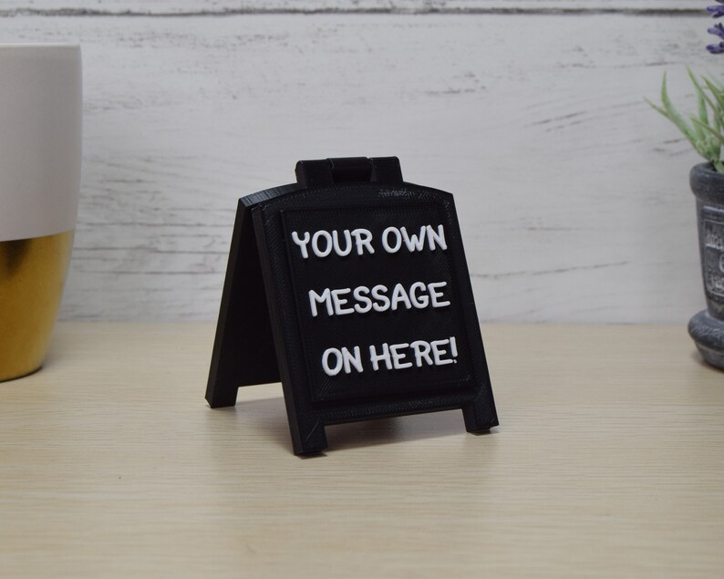 May include: A small, black chalkboard-style sign with white lettering that reads "YOUR OWN MESSAGE ON HERE!". The sign is in an A-frame style, suitable for tabletop display. The background includes a white and gold mug and a small potted plant.