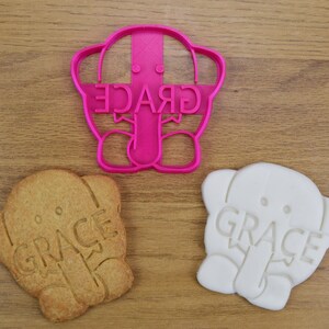 May include: Pink plastic cookie cutter shaped like an elephant with the word "GRACE" written on it. Two cookies, one brown and one white, are also shown, both with the same elephant design and the word "GRACE" imprinted on them.