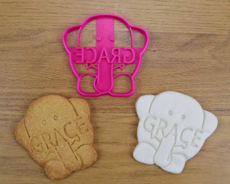May include: Pink plastic cookie cutter shaped like an elephant with the word "GRACE" written on it. Two cookies, one brown and one white, are also shown, both with the same elephant design and the word "GRACE" imprinted on them.