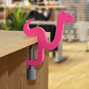 May include: A pink plastic snake clamp with a screw mechanism. The clamp is attached to a wooden surface.