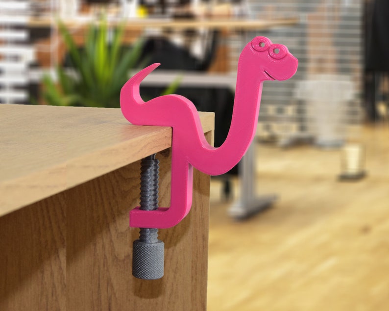 May include: A pink plastic snake clamp with a screw mechanism. The clamp is attached to a wooden surface.