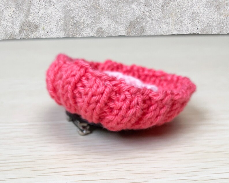 May include: A pink knitted flower brooch with a silver clasp. The flower is made with a textured knit pattern.