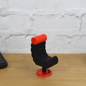 May include: A black and red 3D printed miniature chair with a red padded headrest and a black base. The chair is on a wooden surface and has the word "Henry" printed on the base.