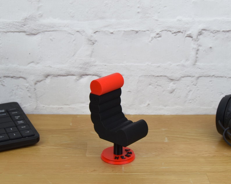 May include: A black and red 3D printed miniature chair with a red padded headrest and a black base. The chair is on a wooden surface and has the word "Henry" printed on the base.