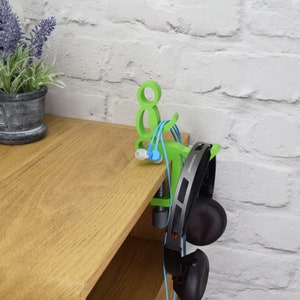 May include: Green 3D printed headphone stand with a clamp to attach to a shelf or desk. The stand has a small figure holding the headphones. The stand is holding a pair of black headphones and a pair of blue and green earbuds.