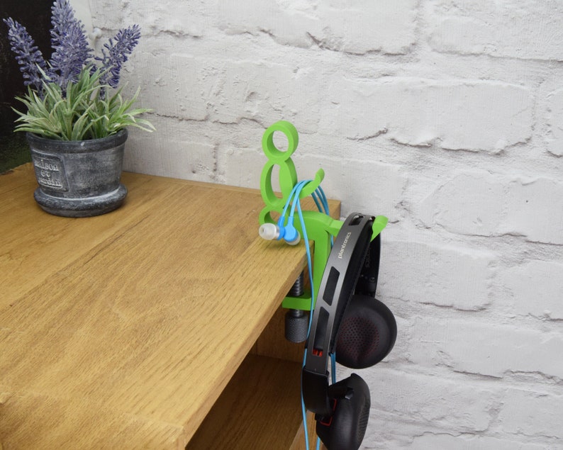 May include: Green 3D printed headphone stand with a clamp to attach to a shelf or desk. The stand has a small figure holding the headphones. The stand is holding a pair of black headphones and a pair of blue and green earbuds.