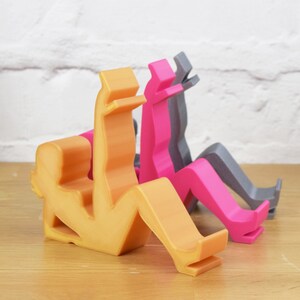 May include: Three phone stands made of plastic in the shape of a person sitting down. The stands are in orange, pink, and gray.