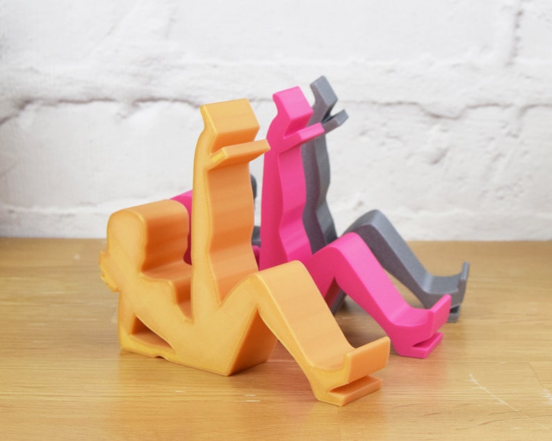 May include: Three phone stands made of plastic in the shape of a person sitting down. The stands are in orange, pink, and gray.