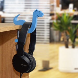 May include: A blue plastic headphone holder shaped like a cartoon snake with a clamp to attach to a desk or shelf. A pair of black Sony headphones are hanging from the holder.