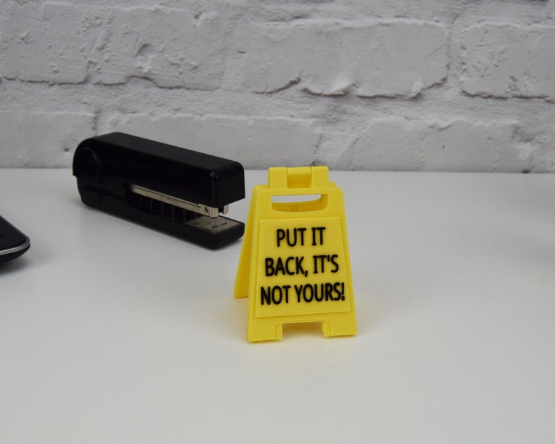 May include: A yellow plastic sign with the text "PUT IT BACK, IT'S NOT YOURS!" stands on a white surface. A black stapler is in the background. The sign is a miniature version of a caution sign.