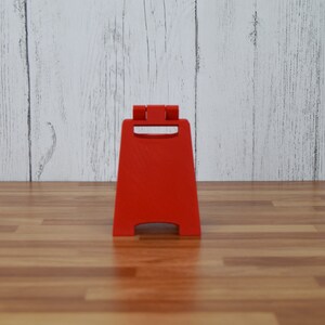 May include: A bright red, freestanding caution sign with a handle at the top. The sign is made of plastic and is designed to stand on its own. The background is a light wood-paneled wall and a wooden floor.