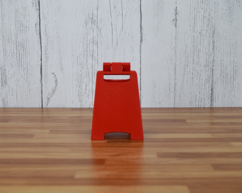May include: A bright red, freestanding caution sign with a handle at the top. The sign is made of plastic and is designed to stand on its own. The background is a light wood-paneled wall and a wooden floor.