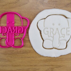 May include: Pink plastic cookie cutter shaped like an elephant with the name "GRACE" written across the front. The cutter is shown next to a white cookie dough imprint of the cutter.
