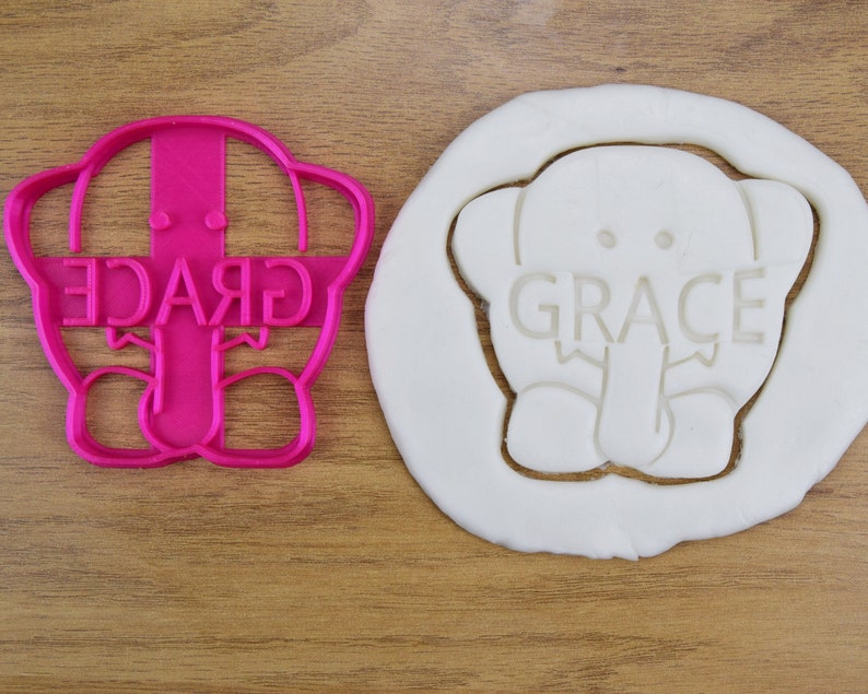 May include: Pink plastic cookie cutter shaped like an elephant with the name "GRACE" written across the front. The cutter is shown next to a white cookie dough imprint of the cutter.