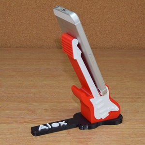 Puede incluir: Un soporte para tel&eacute;fono de color rojo y blanco con forma de guitarra y una base negra. La base tiene el nombre "Alex" impreso.