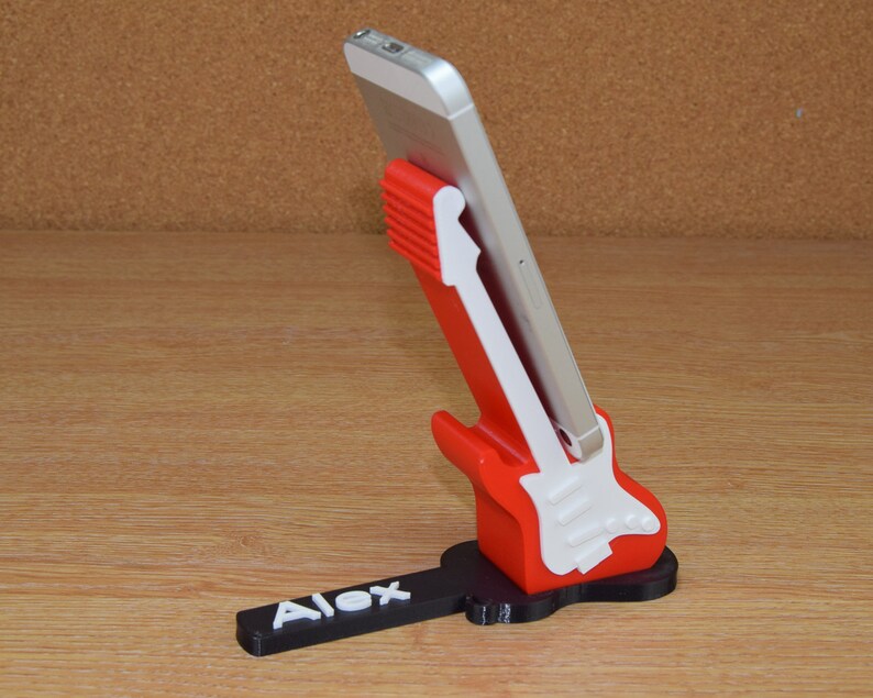 Puede incluir: Un soporte para tel&eacute;fono de color rojo y blanco con forma de guitarra y una base negra. La base tiene el nombre "Alex" impreso.
