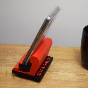 May include: A red phone stand with the words "SOFA SALLY" printed on the side. A smartphone is propped up in the stand.