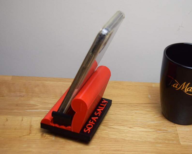May include: A red phone stand with the words "SOFA SALLY" printed on the side. A smartphone is propped up in the stand.