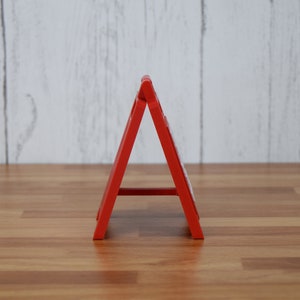 May include: A bright red, A-frame sign, likely for display. The sign is made of a solid material and stands on a wooden surface. The background is a light-colored, wooden wall. The sign is angled, creating a triangular shape.