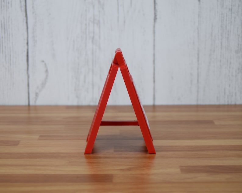 May include: A bright red, A-frame sign, likely for display. The sign is made of a solid material and stands on a wooden surface. The background is a light-colored, wooden wall. The sign is angled, creating a triangular shape.