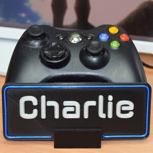 Pu&ograve; includere: Un controller Xbox nero &egrave; posizionato su un supporto nero con un bordo blu. Il supporto ha il nome "Charlie" stampato in bianco sul davanti.