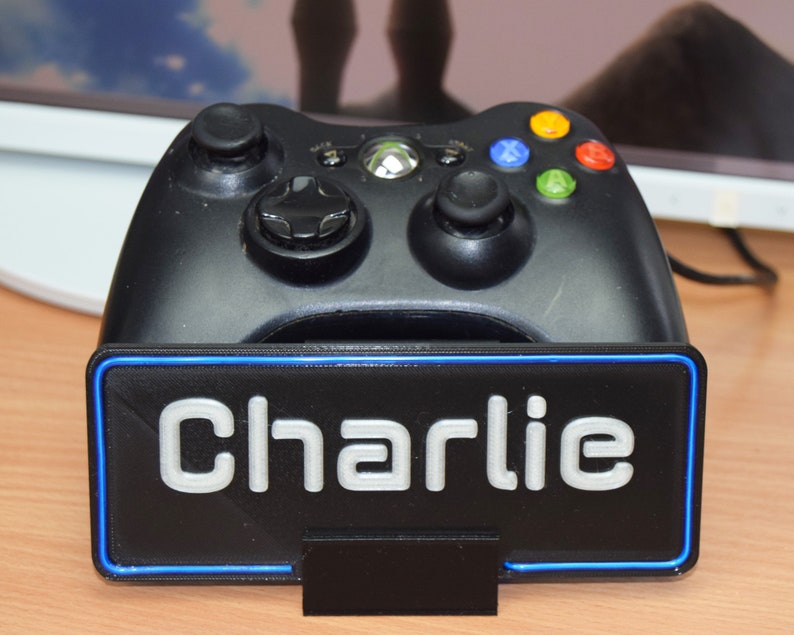 Pu&ograve; includere: Un controller Xbox nero &egrave; posizionato su un supporto nero con un bordo blu. Il supporto ha il nome "Charlie" stampato in bianco sul davanti.