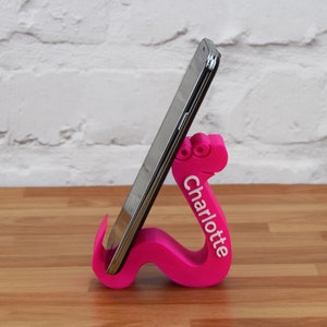 May include: A pink 3D printed phone stand shaped like a worm with the name "Charlotte" printed on the side. A black smartphone is resting in the stand.