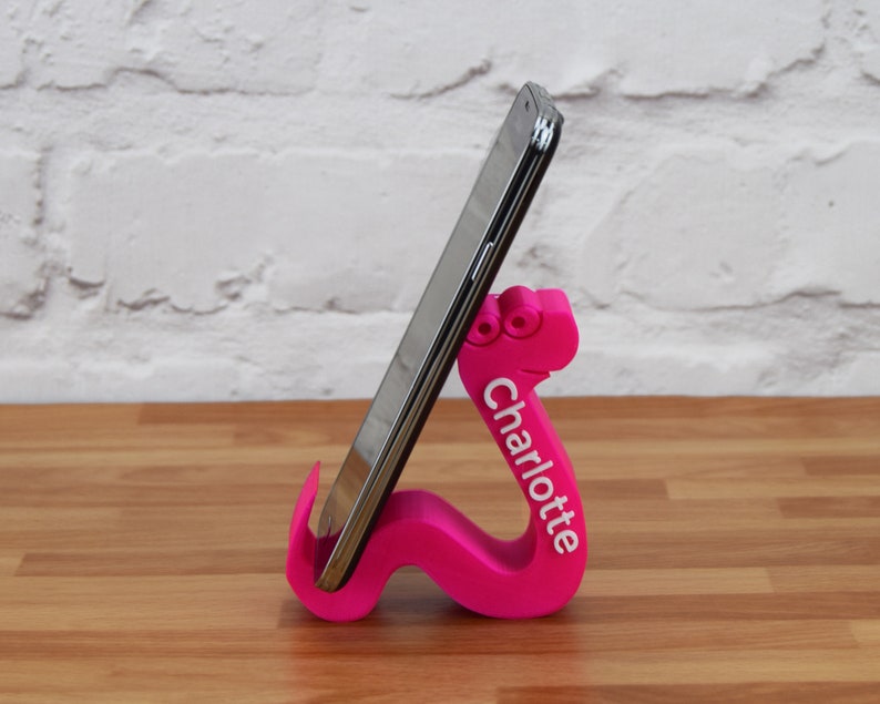 May include: A pink 3D printed phone stand shaped like a worm with the name "Charlotte" printed on the side. A black smartphone is resting in the stand.