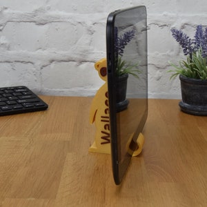 May include: A wooden tablet stand shaped like a meerkat with the word "Wallace" printed on the side. The stand is holding a black tablet in portrait mode.