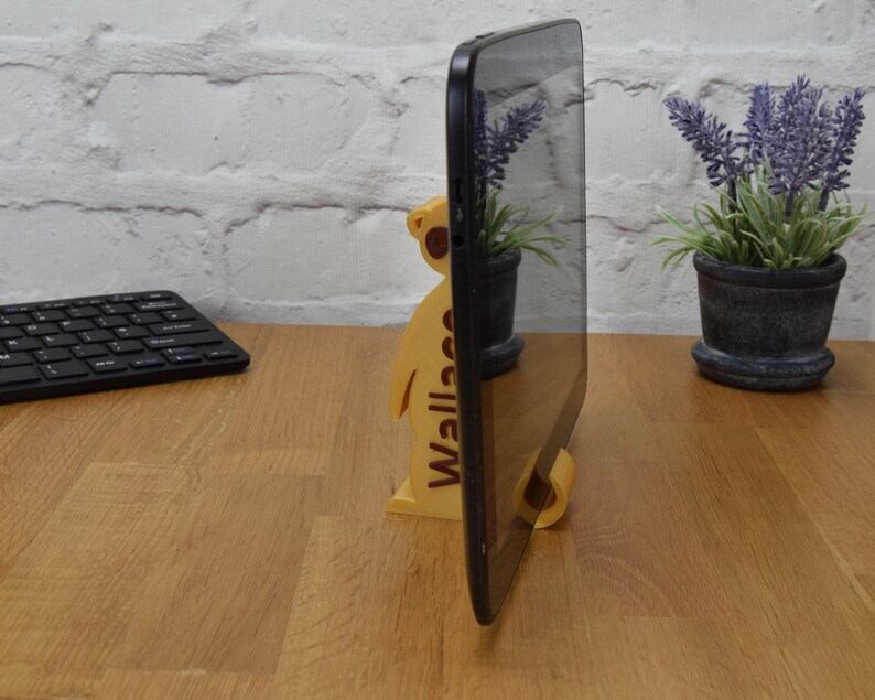 May include: A wooden tablet stand shaped like a meerkat with the word "Wallace" printed on the side. The stand is holding a black tablet in portrait mode.