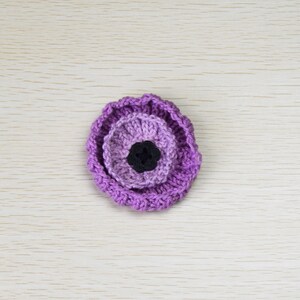 May include: A purple crocheted flower with a black center. The flower is made of multiple layers of crocheted fabric.
