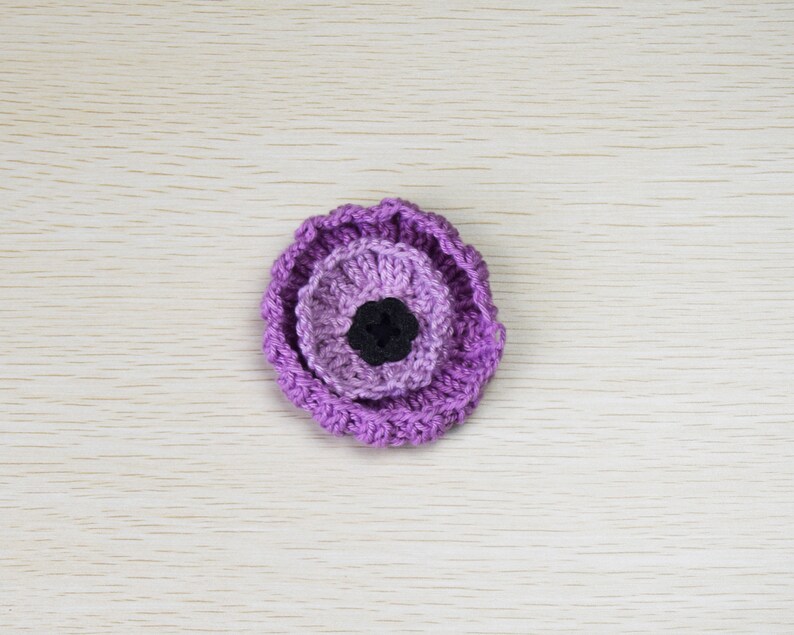 May include: A purple crocheted flower with a black center. The flower is made of multiple layers of crocheted fabric.