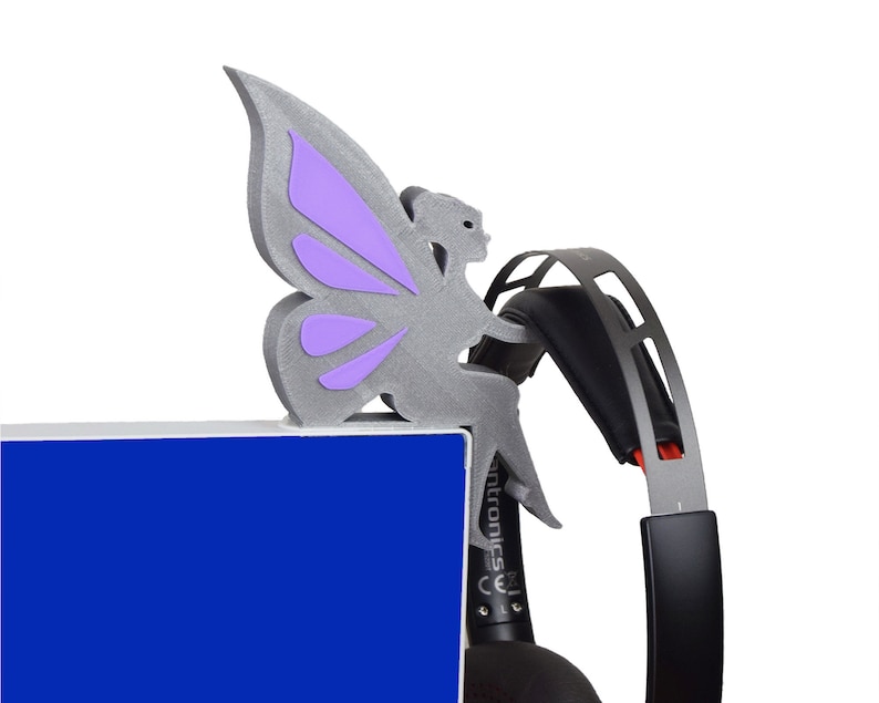 K&ouml;nnte beinhalten: Eine graue, 3D-gedruckte Feenfigur mit lila Fl&uuml;geln, die auf einem schwarzen Headset sitzt. Die Fee sitzt auf einer blauen Oberfl&auml;che.