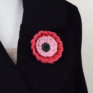 May include: A pink and red crocheted flower brooch with a black button center.