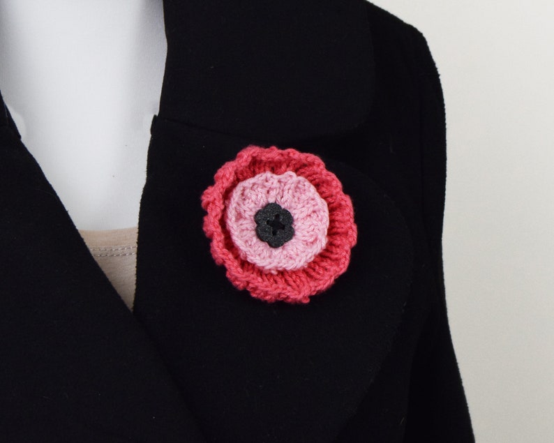 May include: A pink and red crocheted flower brooch with a black button center.