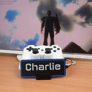 Pu&ograve; includere: Un controller per videogiochi bianco con pulsanti neri &egrave; visualizzato su un supporto con una luce blu. Il supporto ha uno sfondo nero con la parola "Charlie" stampata in bianco.