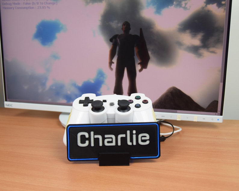 Pu&ograve; includere: Un controller per videogiochi bianco con pulsanti neri &egrave; visualizzato su un supporto con una luce blu. Il supporto ha uno sfondo nero con la parola "Charlie" stampata in bianco.