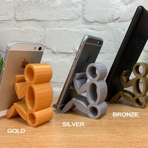 May include: Three 3D printed phone stands in gold, silver, and bronze. Each stand is shaped like a person holding a phone.