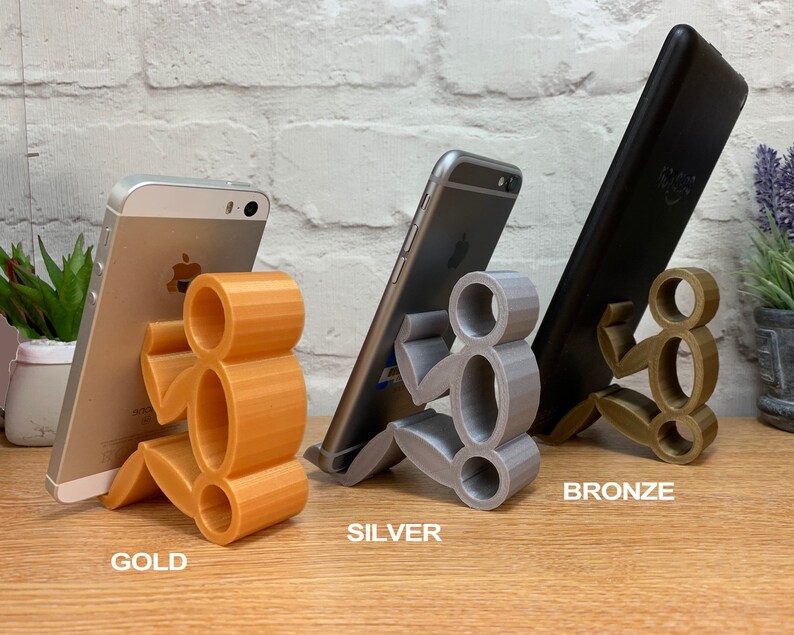 May include: Three 3D printed phone stands in gold, silver, and bronze. Each stand is shaped like a person holding a phone.