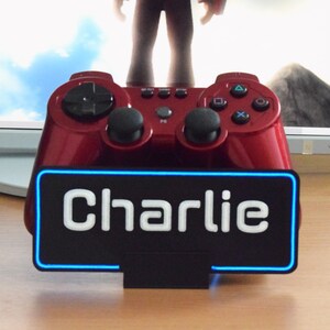 Pu&ograve; includere: Un controller per videogiochi rosso con una base nera e un cartello al neon blu che dice "Charlie".