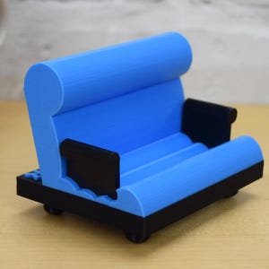 May include: A blue and black 3D printed phone stand shaped like a couch. The stand has a rounded back and armrests, and a flat surface for holding a phone.