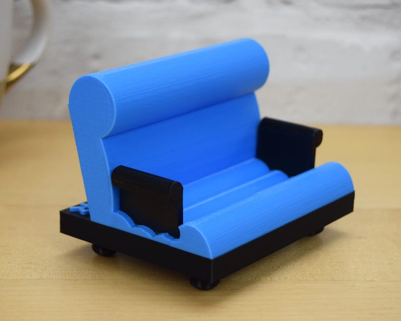 May include: A blue and black 3D printed phone stand shaped like a couch. The stand has a rounded back and armrests, and a flat surface for holding a phone.
