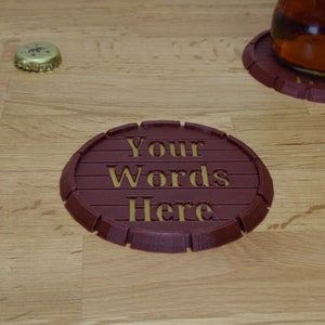 May include: A brown, round, 3D printed coaster with the words "Your Words Here" in gold lettering.