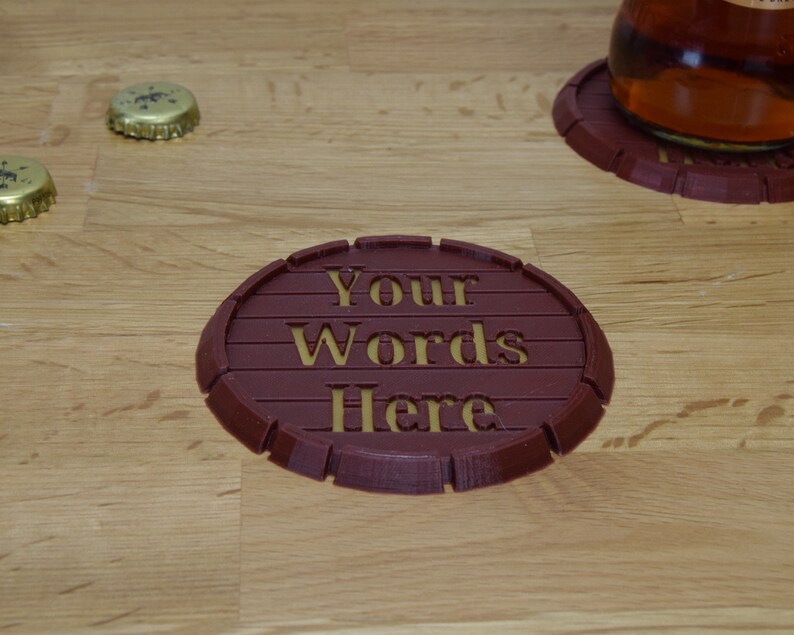 May include: A brown, round, 3D printed coaster with the words "Your Words Here" in gold lettering.