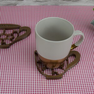 May include: A white ceramic mug with a copper-colored base and gold handle sits on a brown, cup-shaped coaster. The coaster has a caffeine molecule design and the word "Caffeine" on it. Another coaster is to the left.