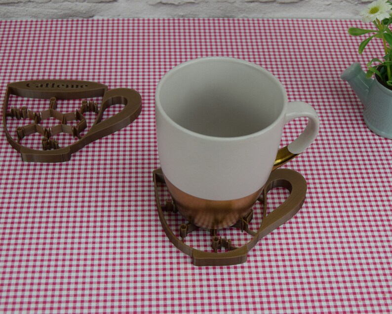 May include: A white ceramic mug with a copper-colored base and gold handle sits on a brown, cup-shaped coaster. The coaster has a caffeine molecule design and the word "Caffeine" on it. Another coaster is to the left.