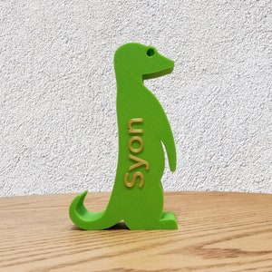 K&ouml;nnte beinhalten: Eine gr&uuml;ne Plastikfigur eines Erdm&auml;nnchens, das auf einer Holzoberfl&auml;che steht. Das Erdm&auml;nnchen schaut nach rechts und hat das Wort "S'yon" in goldenen Buchstaben auf der Seite geschrieben.